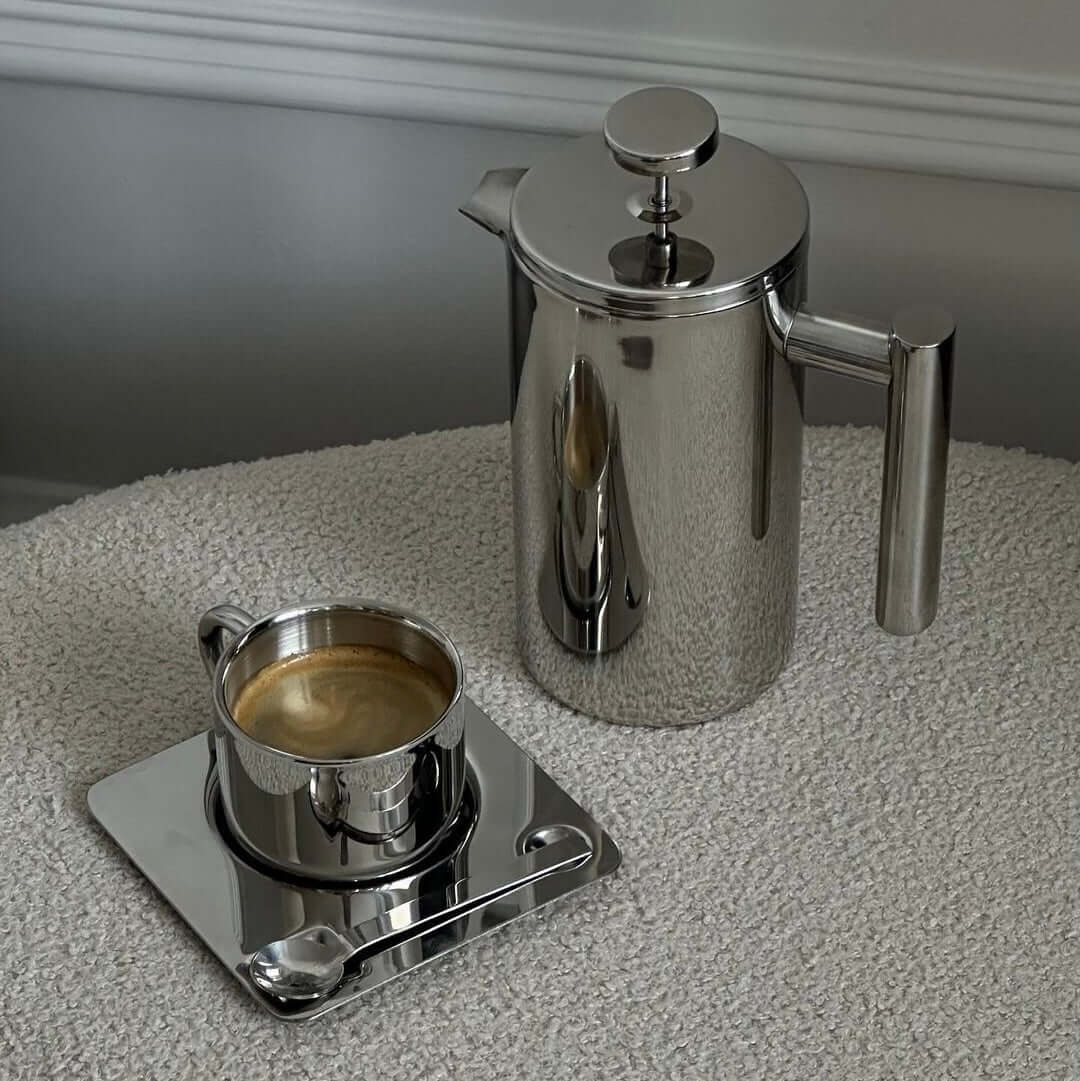 Stainless steel French press coffee pot with mirror finish on kitchen counter with cups and bottle.