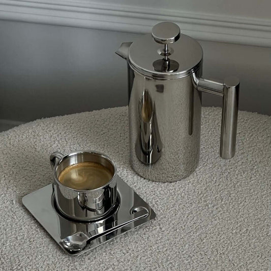 Stainless steel French press coffee pot with mirror finish on kitchen counter with cups and bottle.