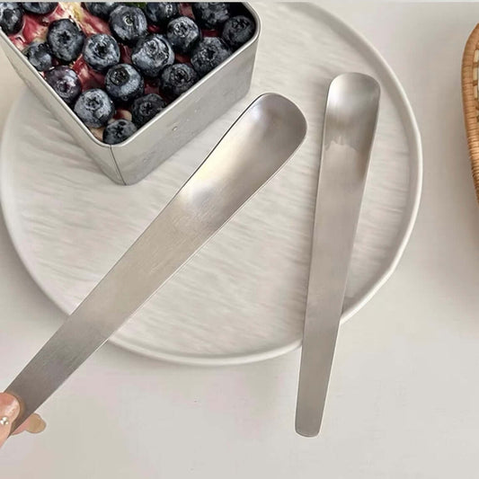 Minimalist dessert spoon with measurements of 12.2cm length and 1.5cm width displayed beside a scalloped plate.